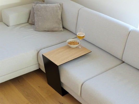 Couchtisch aus Metall, Farbe Scwharz und auf der Oberseite ist ein Eiche Furnier drauf geklebt. In Trapezform und U-Form der über ein Sofa geschoben ist. Auf dem kleinen Couchtisch steht ein Weinglas befüllt mit Bier und daneben eine Schalle mit erdnüssen.