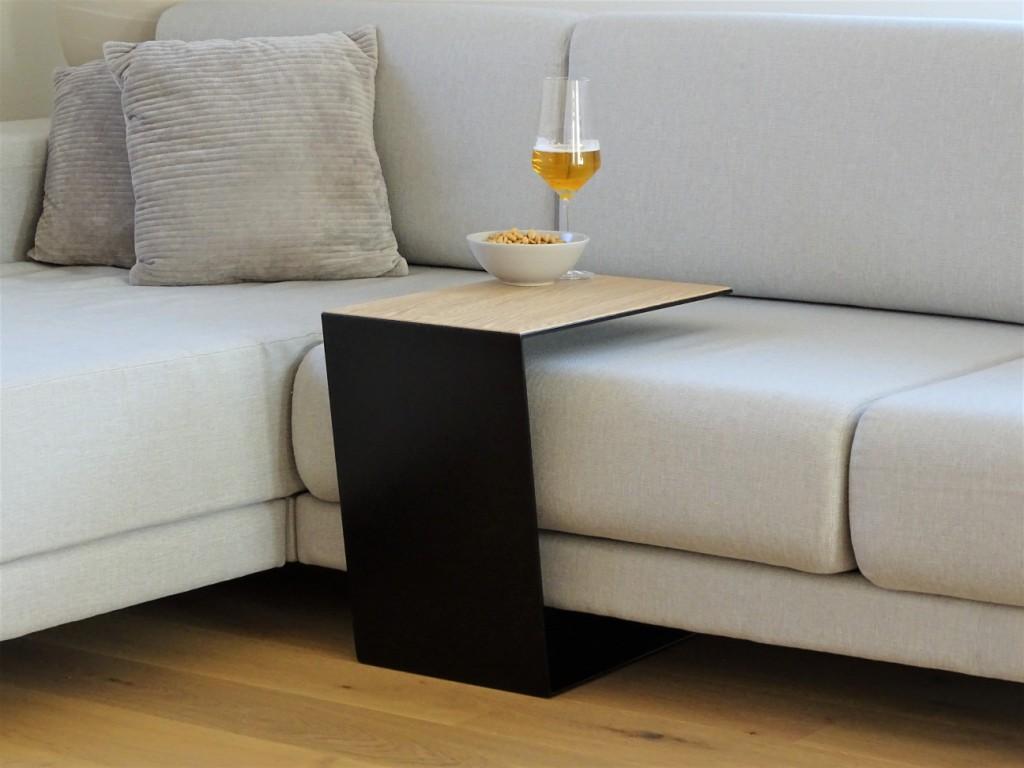 Couchtisch aus Metall, Farbe Scwharz und auf der Oberseite ist ein Eiche Furnier drauf geklebt. In Trapezform und U-Form der über ein Sofa geschoben ist. Auf dem kleinen Couchtisch steht ein Weinglas befüllt mit Bier und daneben eine Schalle mit erdnüssen.