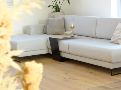 Couchtisch aus Metall, Farbe Scwharz und auf der Oberseite ist ein Eiche Furnier drauf geklebt. In Trapezform und U-Form der über ein Sofa geschoben ist. Auf dem kleinen Couchtisch steht ein Weinglas befüllt mit Bier und daneben eine Schalle mit erdnüssen.