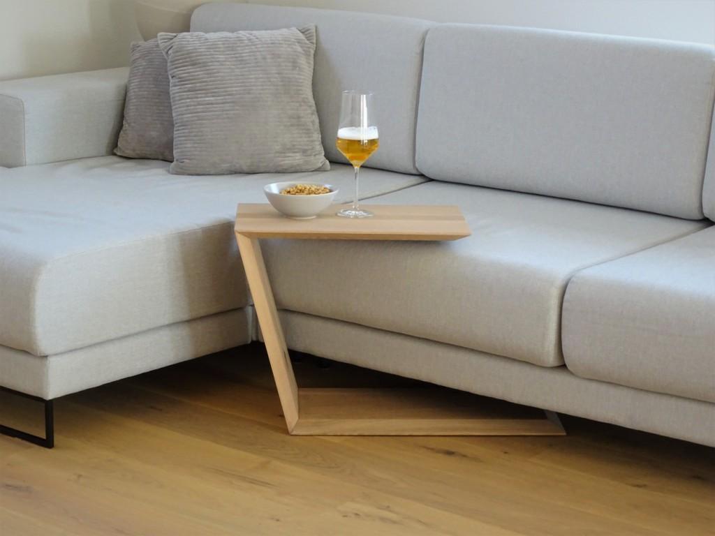 Couchtisch in Trapezform und U-Form der über ein Sofa geschoben ist. Auf dem kleinen Couchtisch steht ein Weinglas befüllt mit Bier und daneben eine Schalle mit erdnüssen.