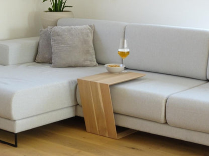 Couchtisch in Trapezform und U-Form der über ein Sofa geschoben ist. Auf dem kleinen Couchtisch steht ein Weinglas befüllt mit Bier und daneben eine Schalle mit erdnüssen.